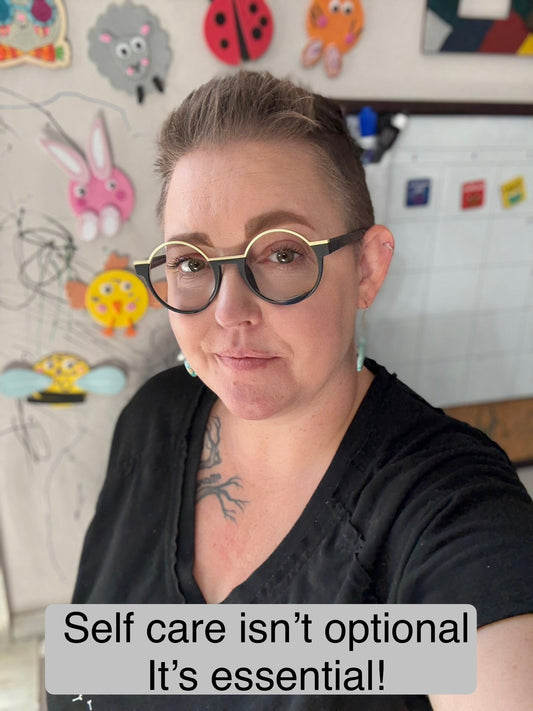 Person with short hair and glasses takes a selfie in front of a colorful craft wall. Caption reads: “Self care isn’t optional. It’s essential!”