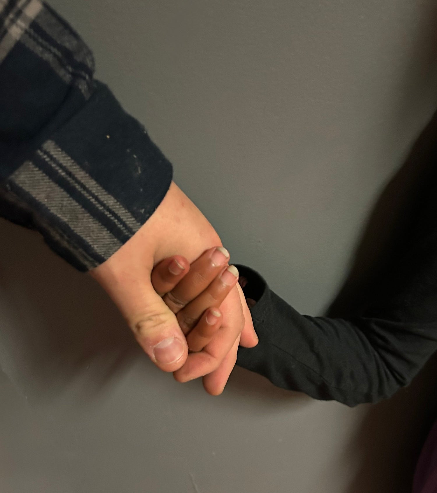 A close-up of a white adult hand gently holding the hand of a Black child, fingers intertwined, against a gray wall background.